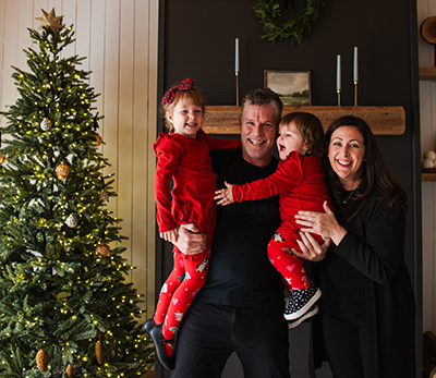 Ashley, her partner, and two children dressed in holiday reds and blacks beside their Christmas tree