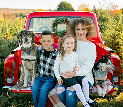 Karley and her two children and dog, sitting in the back of a red pickup truck in a Christmas tree orchard.